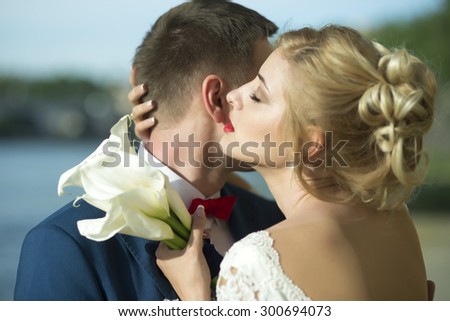 Beautiful young wedding couple of man in blue jacket and red tie bow embracing blonde woman in white dress holding bouquet of calla flowers standing on sunny outdoor background, horizontal picture