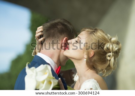 Attractive young wedding couple of man in blue jacket and red tie bow embracing blonde girl in white dress holding posy of calla flowers standing on sunny outdoor background, horizontal picture