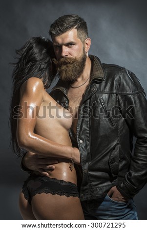 Couple of young undressed woman in panties with soft skin and bare chest embracing unshaven guy with beard in brown leather biker jacket standing on black background, vertical picture