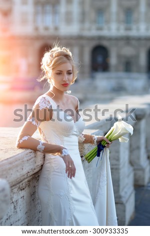 Beautiful sensory young blond bride with stylish hairdo in white wedding dress holding bouquet of calla flowers with blue ribbon standing on stone bridge outdoor sunny day, vertical picture