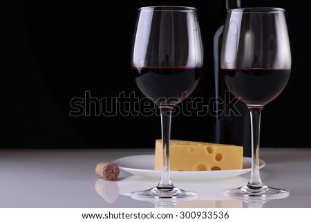 Two glasses of red wine standing with bottle and fresh tasty cheese on plate and cork on white table top on black studio background copyspace, horizontal picture