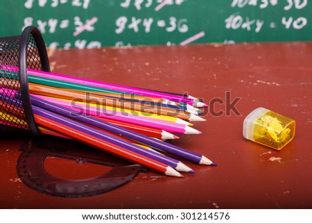 Colorful pencils of red yellow orange violet purple pink green and blue in stationary cup ruler and steel lying on brown school desk on written with white chalk blackboard backgroung on lesson of math