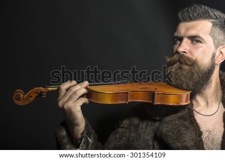 Musical brutal unshaven man with long beard and hendlebar moustache in brown fur coat with collar and chain on chest holding wooden violin standing on black background copyspace, horizontal picture