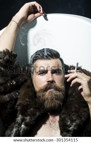 Confident sexy unshaven man with long beard and moustache in brown fur coat with collar and blue jeans lying in white bath tub with raised hand smoking cigarette, vertical picture