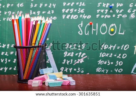 Colorful pencils of red yellow orange violet purple pink green and blue in stationary cup and chalk standing on brown school desk on written blackboard backgroung on lesson of math