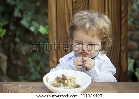 Funny pretty little baby boy with blond curly hair with cute expression on face sitting on wooden chair eating cereal for breakfast from plate outdoor on natural green background, horizontal photo