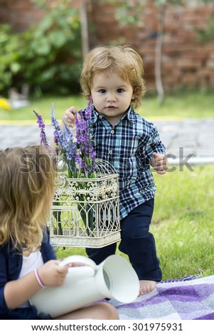 Pretty small boy with blond curly hair in blue checkered shirt and jeans holding white iron cage with purple and violet field flowers giving to girl friend sunny day outdoor, vertical picture