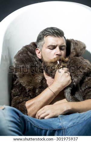 Confident unshaven man with long beard and moustache in brown fur coat with collar and blue jeans lying in white bath tub smoking cigarette, vertical picture
