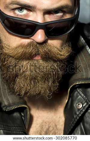 Portrait of sexual brutal unshaved guy with long beard and moustache in brown leather biker jacket and black sunglasses looking forward closeup, vertical picture