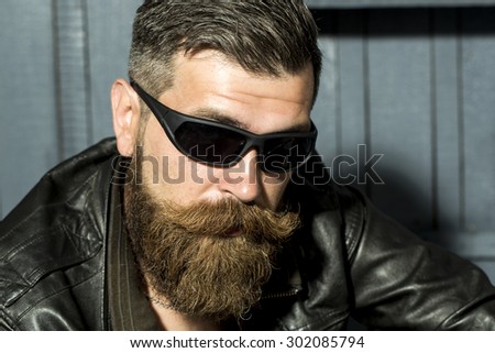 Portrait of handsome brutal unshaved male biker with long beard and moustache in brown leather jacket and black sunglasses sitting on grey wooden background, horizontal picture