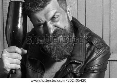 Cool brutal unshaven man with long beard and moustache holding empty glass wine bottle near head sitting leather jacket on wooden background black and white, horizontal picture