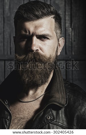 Portrait of beautiful brutal unshaven man with long beard and hendlebar in brown leather jacket and chain looking forward with puckered brows standing on grey wooden background, vertical picture