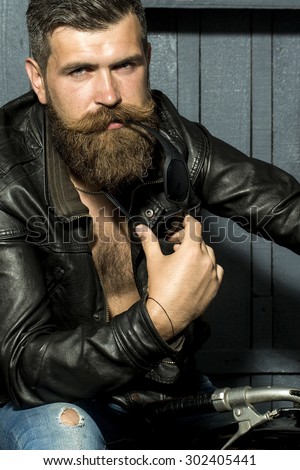 Handsome brutal unshaven sexy male biker in brown leather jacket jeans with sun glasses sitting in garage on motorcycle looking forward on wooden background, vertical picture