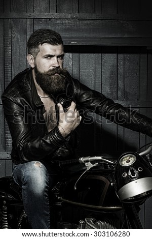 Handsome brutal unshaven sexual male biker in black leather jacket jeans with sun glasses sitting in garage on motor bike looking forward on wooden background, vertical picture