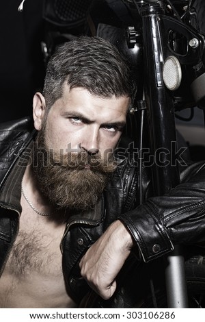Portrait of serious sexy unshaven man with beard and handlebar moustache in black leather biker jacket with bare torso sitting near motorcycle looking forward, vertical picture