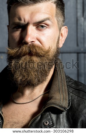 Portrait of sullen brutal unshaven man with long beard and hendlebar in brown leather jacket and chain looking forward with puckered brows standing on grey wooden background, vertical picture