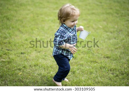 Cute small baby boy with blond curly hair in blue checkered shirt and jeans holding white bucket running on green grass lawn sunny day outdoor copyspace, horizontal picture