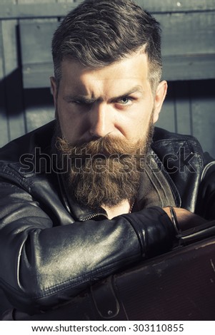 Portrait of sullen unshaven male biker in black leather jacket sitting in garage with big brown old briefcase looking forward on workshop background, vertical picture