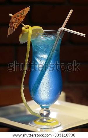 One glass with alcoholic blue lagoon cocktail of liqueur blue curacao vodka sprite yellow lemon citrus fruit and ice cubes with drink straws and umbrella on brick background, vertical photo
