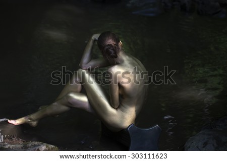 Beautiful naked woman mermaid with sexy painted gold body sitting with back on stony river coast in forest near water sunny day outdoor on natural background, horizontal picture