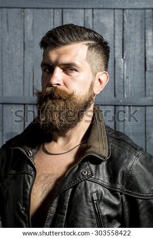 Portrait of attractive brutal unshaven man with long beard and hendlebar in brown leather jacket and chain looking forward standing on grey wooden background, vertical picture