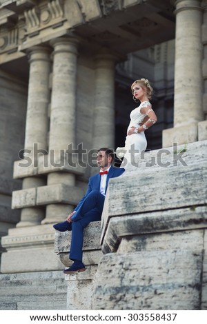 Beautiful young wedding couple of man in blue suit and blond woman in long white dress sitting in entrance of stone building of Corte di Cassazione in Rome Italy sunny day outdoor, vertical picture