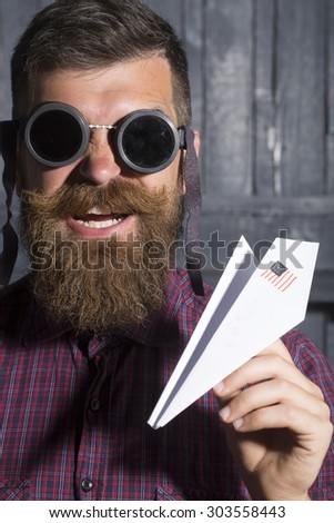 Poprtrait of young emotional man in purple checkered shirt and aviator glasses standing in garage holding white paper plane with usa flag looking forward on workshop background, vertical picture