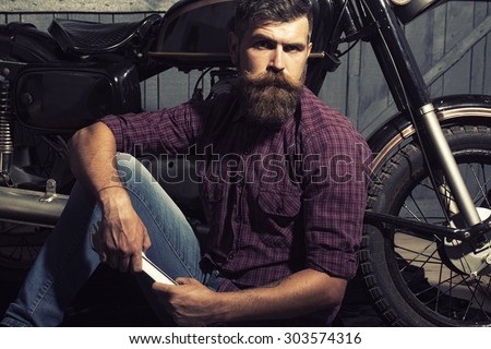 Attractive unshaven male biker in purple checkered shirt and jeans sitting near motorcycle in garage holding metallized iron spanner looking forward on workshop background, horizontal picture