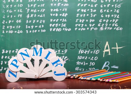 Colorful pencils of red yellow orange violet purple pink green and blue near stationary cup and fan english alphabet standing on brown school desk on written with white chalk blackboard on math