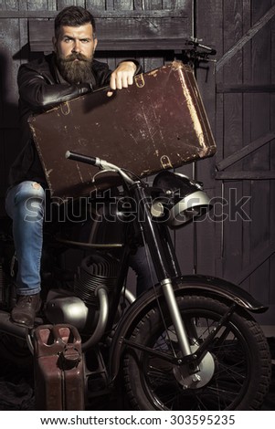 Handsome unshaven male biker in leather jacket sitting near motorcycle in garage with big brown old briefcase and rusty fuel can looking forward on workshop background, vertical picture