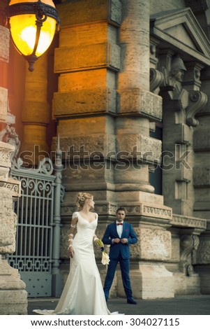 Beautiful stylish young wedding couple of blonde girl in long white dress with calla flower bunch looking at man in blue suit standing near stone building with illuminated street lamps, vertical photo