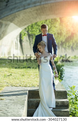 Beautiful young tender wedding couple of blonde woman in long white dress with calla flower bunch and man in blue suit with red bow tie standing under stone bridge on stairs in sunbeam, vertical photo