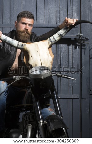 Attractive unshaven male biker in leather jacket sitting on motorcycle in garage with big bone skull antlers of stuffed animal looking forward on wooden wall background, vertical picture