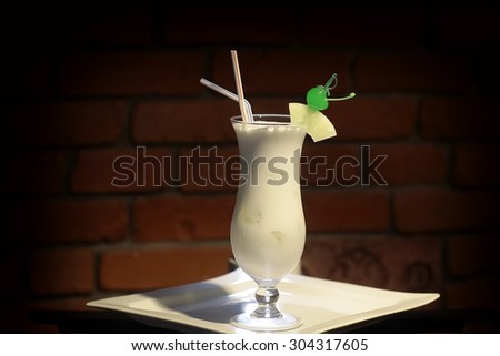 Glass with alcoholic pina colada cocktail of light rum coir coconut milk crushed ice frappe pineapple juice and slice green cherry and drink straws on white plate on brick background, horizontal photo