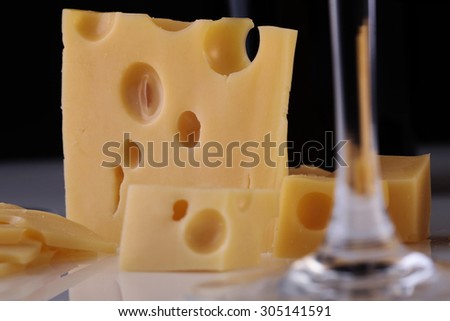 Closeup of fresh yellow delicious delicatessen dairy product of cheese slices near stem of crystal glass wine goblet standing on white table top on black background, horizontal picture
