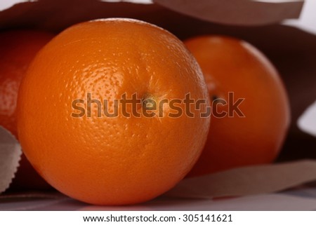 Closeup of fresh juisy bright tasty whole orange in brown paper packet with heap of citrus fruit lying on white reflecting table top in studio, horizontal picture