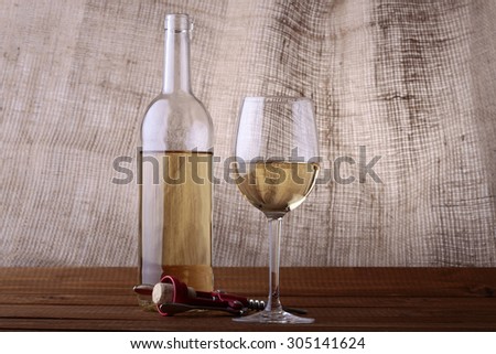 One glass open bottle and bocals with white wine standing on brown wooden table top near red plastic and metal corkscrew with cork on burlap background, horizontal picture