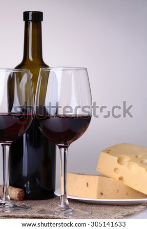 Fresh yellow delicious delicatessen dairy product of cheese slices on plate near crystal glass goblets and bottle with red wine standing on white table top on grey background, vertical picture