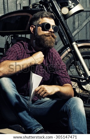 Serious unshaven male biker in purple checkered shirt and glasses sitting near motorcycle in garage holding white paper plane looking forward on workshop background, vertical picture