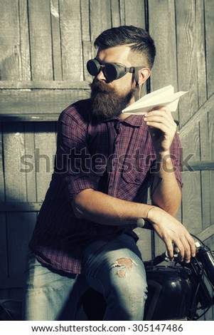 Handsome unshaven male biker in purple checkered shirt and glasses standing near motorbike in garage holding white paper plane looking forward on workshop background, vertical picture