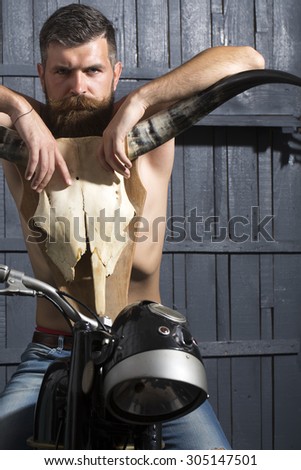 Handsome sexy undressed unshaven male biker in jeans sitting on motorcycle in garage with big bone skull antlers of stuffed animal looking forward on wooden wall background, vertical picture