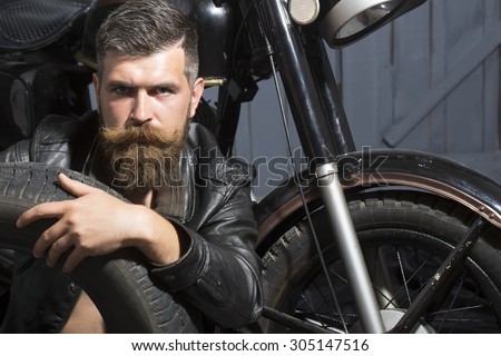 Handsome unshaven male biker in leather jacket sitting near motorcycle in garage with big black rubber spare wheel looking forward on wooden wall background, horizontal picture