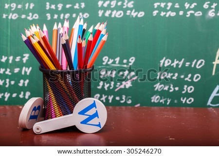 Colorful pencils of red yellow orange violet purple pink green and blue in stationary cup ruler and fan english alphabet standing on brown school desk on written with white chalk blackboard on math