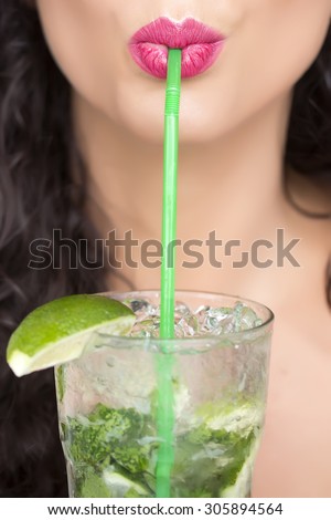 Closeup of sexual pretty brunette female face of girl with curly hair and pink lips drinking alcoholic mojito cocktail from mint soda light rum and lime with straw, vertical picture