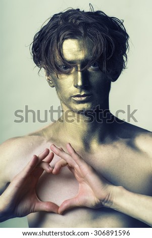 One young fashionable painted man model with golden bodyart on face and stylish hairdo holding hands in shape of heart on bare chest standing in studio on white background, vertical picture