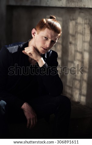 Cute young attractive man with red hair in ponytail sitting sunny day outdoor in black jacket thinking and looking forward on stone background, vertical picture