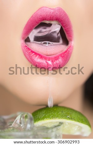 Closeup of sexy open female mouth of attractive young woman with bright pink lips holding cold fresh crystal ice cube from glass of alcoholic mojito cocktail with lime, vertical picture