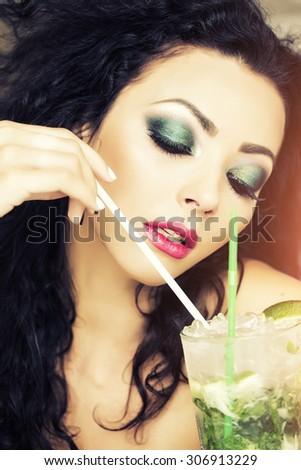 Portrait of alluring pretty brunette woman with curly hair and bright makeup with alcoholic mojito cocktail from mint soda light rum ice cubes and lime with straw, vertical picture