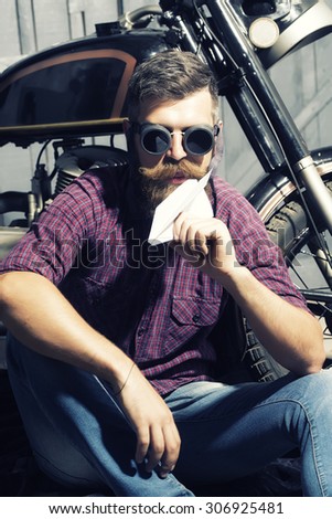 Cool unshaven male biker in purple checkered shirt and glasses sitting near motorcycle in garage holding white paper plane looking forward on workshop background, vertical picture