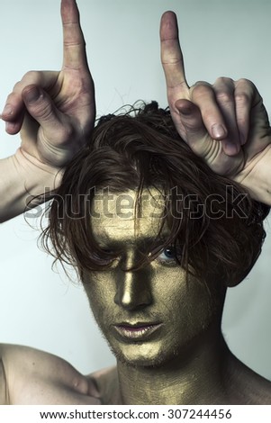Portrait of young fashionable painted man model with golden bodyart on face and stylish hairdo holding hands as antlers looking forward, vertical picture